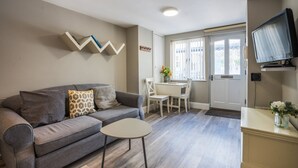A living room with a grey sofa, a small round table, a TV on a stand, and a dining area with a white table and chairs. - Apartment 32 Trinity Mews - sleeps 3 guests  in 1 bedroom (Torquay)