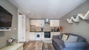 A modern kitchen with white cabinets, a built-in oven, and a washing machine. There is a grey sofa with a patterned pillow and a small white table in the living area. - Apartment 32 Trinity Mews - sleeps 3 guests  in 1 bedroom (Torquay)