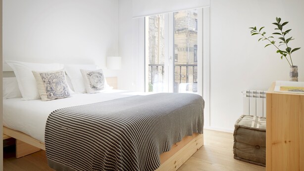 A modern bedroom with a large bed, a view of the city through a window, and a small wooden side table with a plant. - Santa María by FeelFree Rentals (San Sebastián)