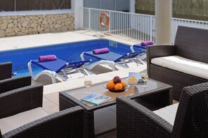 Uma área à beira da piscina com espreguiçadeiras, uma mesa com um livro, uma fruteira e copos.