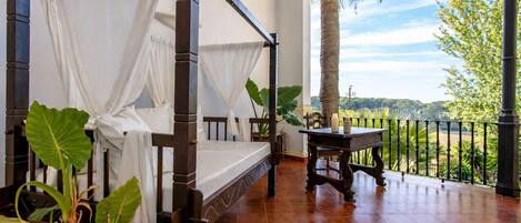Una cama con dosel adornada con cortinas blancas, una mesa de madera con velas y vistas a un paisaje exuberante a través de la barandilla de un balcón.
