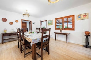 Un comedor con una mesa de madera, sillas y una araña. Hay cuadros en las paredes y una ventana con vista a follaje.
