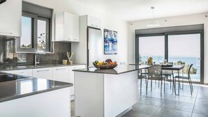 A modern kitchen with white cabinetry, a central island with a fruit bowl, and a dining area with a view of the sea.