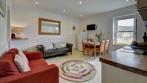 Una sala moderna con un sofá rojo, un sofá de cuero, una mesa de comedor con sillas, un televisor montado en la pared y una cocina con ventana.
