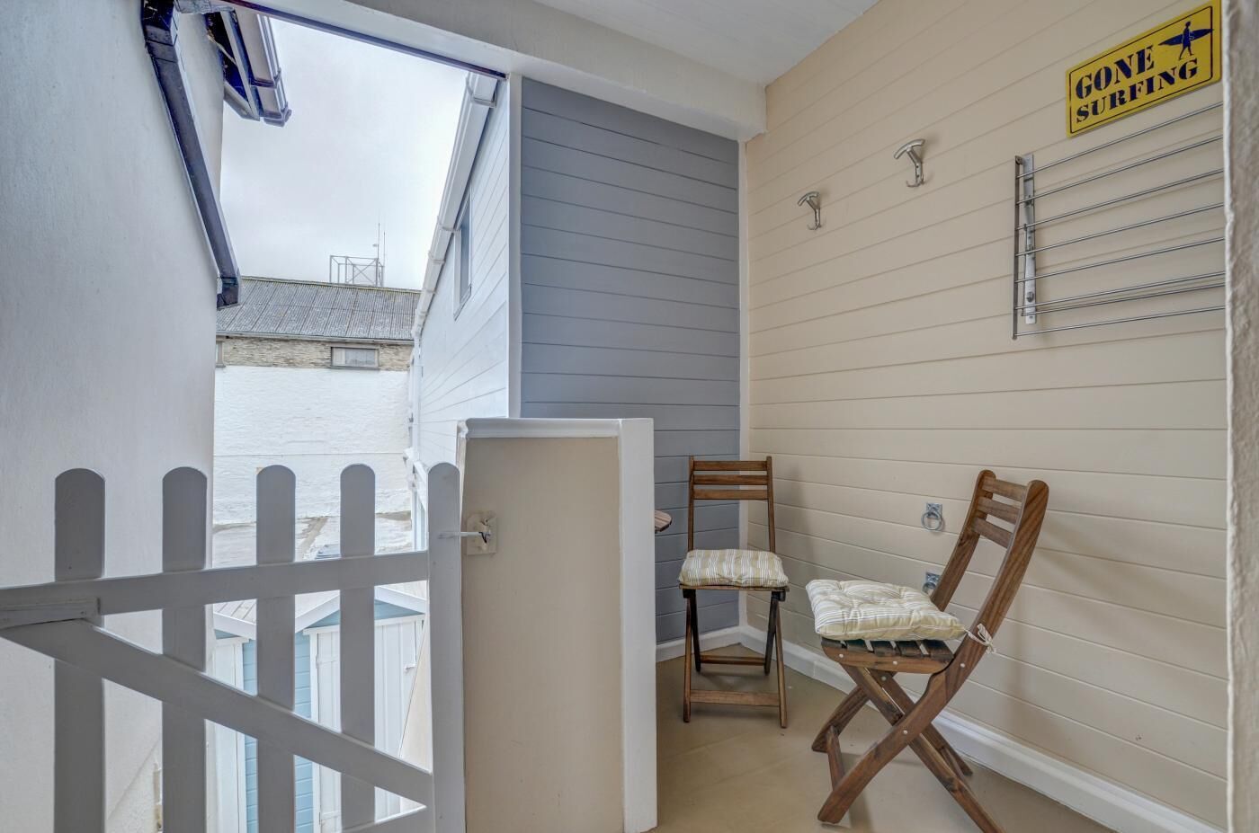 A balcony with a white picket fence, a wooden chair, a small table, and a 'GONE SURFING' sign on the wall.
