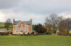 Une grande maison en briques, dotée d’un toit à pignon, entourée d’un champ verdoyant et d’arbres.