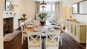 A dining room with a wooden table set for a meal, white chairs, a fireplace, and a view through the window. - Upper Mowley -  a spacious farmhouse sleeping sixteen guests (Kington)