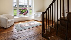 A room with a large window, a wooden floor, a white chair, a white armchair, and a staircase with a wooden railing. - Upper Mowley -  a spacious farmhouse sleeping sixteen guests (Kington)