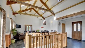Un vaste espace de vie doté d’un plafond haut, de poutres en bois, d’un coin salon avec un canapé et des fauteuils, d’un téléviseur et d’une porte à panneaux de verre.