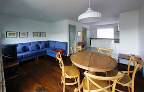A living room with a blue sofa, wooden dining table, and chairs, a television, and framed pictures on the wall. - Higher O'Connell 2 Kinsale, Co. Cork (Kinsale)