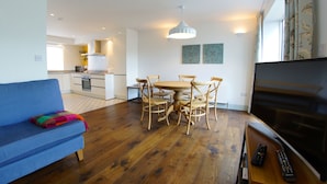 A modern living room with a blue sofa, wooden dining table, and chairs. A TV is on a wooden stand, and a remote control is on the stand. - Higher O'Connell 2 Kinsale, Co. Cork (Kinsale)
