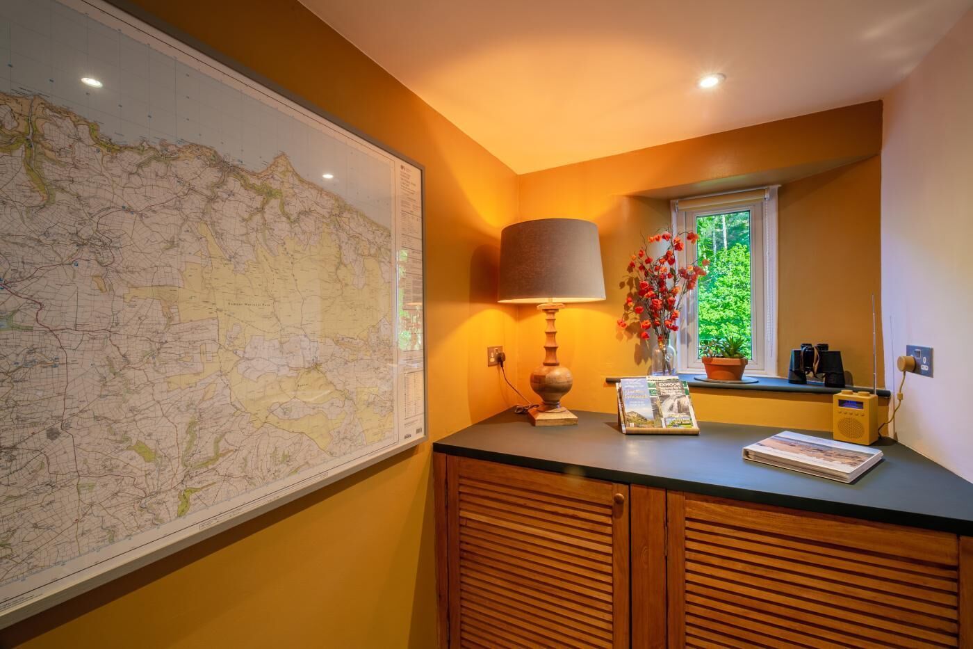 Une pièce avec une grande carte accrochée au mur, une lampe, une fenêtre donnant sur un paysage verdoyant et une petite table sur laquelle est posé un vase de fleurs.