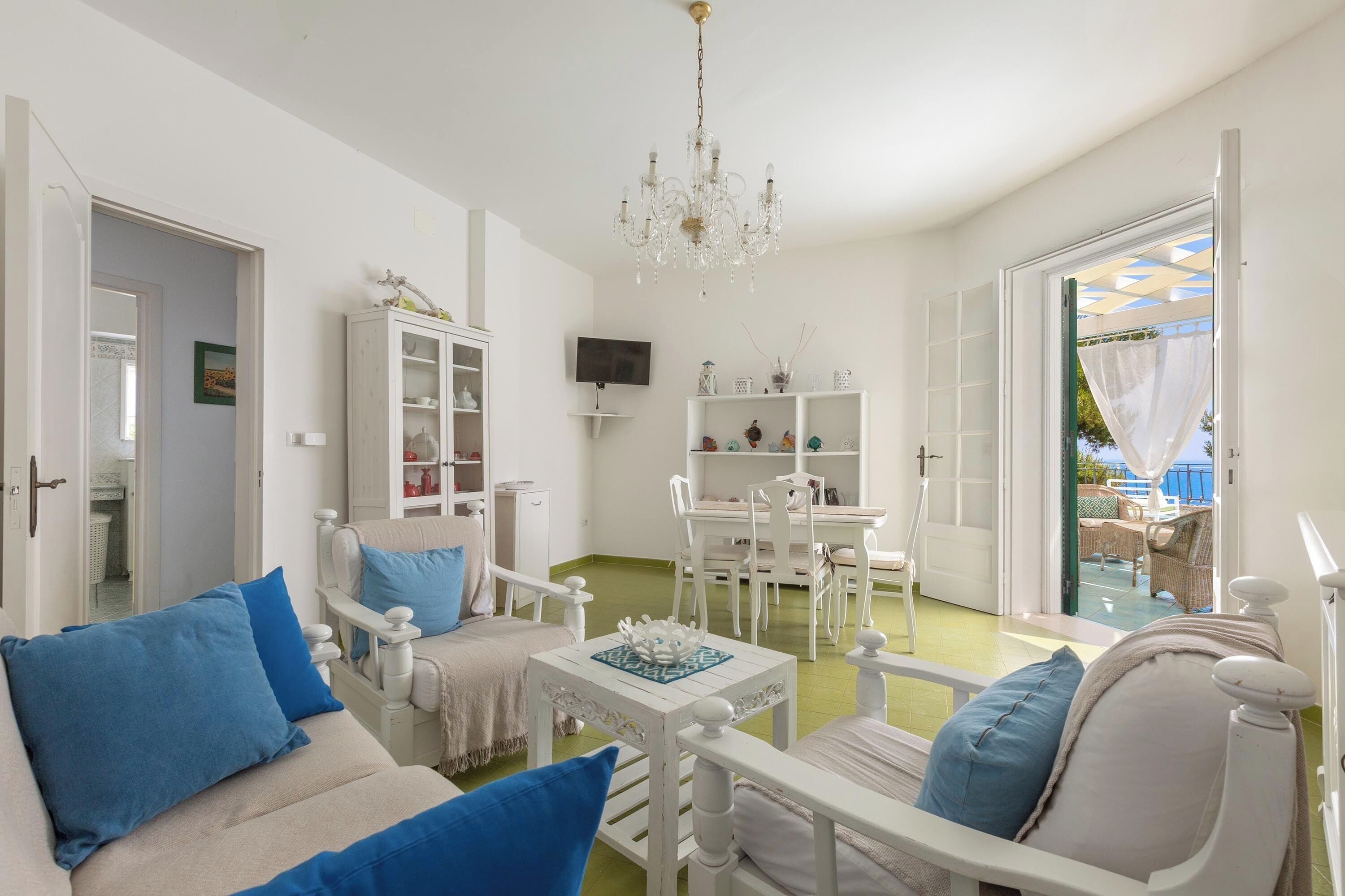 A bright living room with white furniture, a chandelier, and a view of the outside.