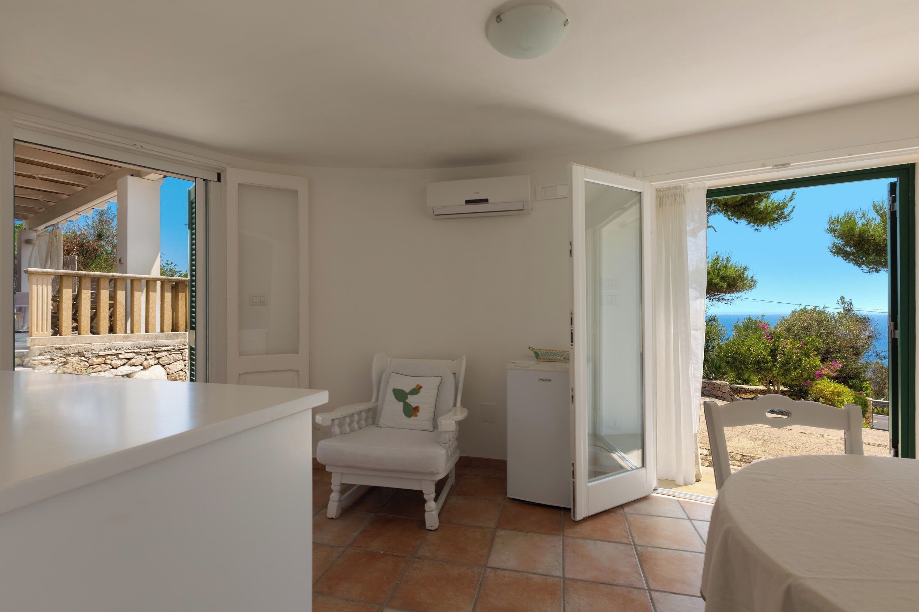 Una habitación con una silla blanca, una mesita y vista al mar a través de una puerta abierta.