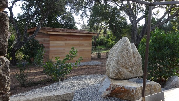 Una terraza de madera con un banco y una mesa, rodeada de rocas y una pequeña construcción de madera.