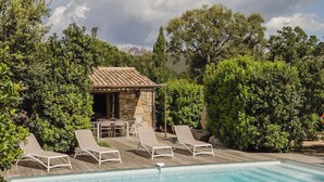 Un área de piscina con sillones de descanso, una cocina al aire libre de piedra y abundante vegetación.
