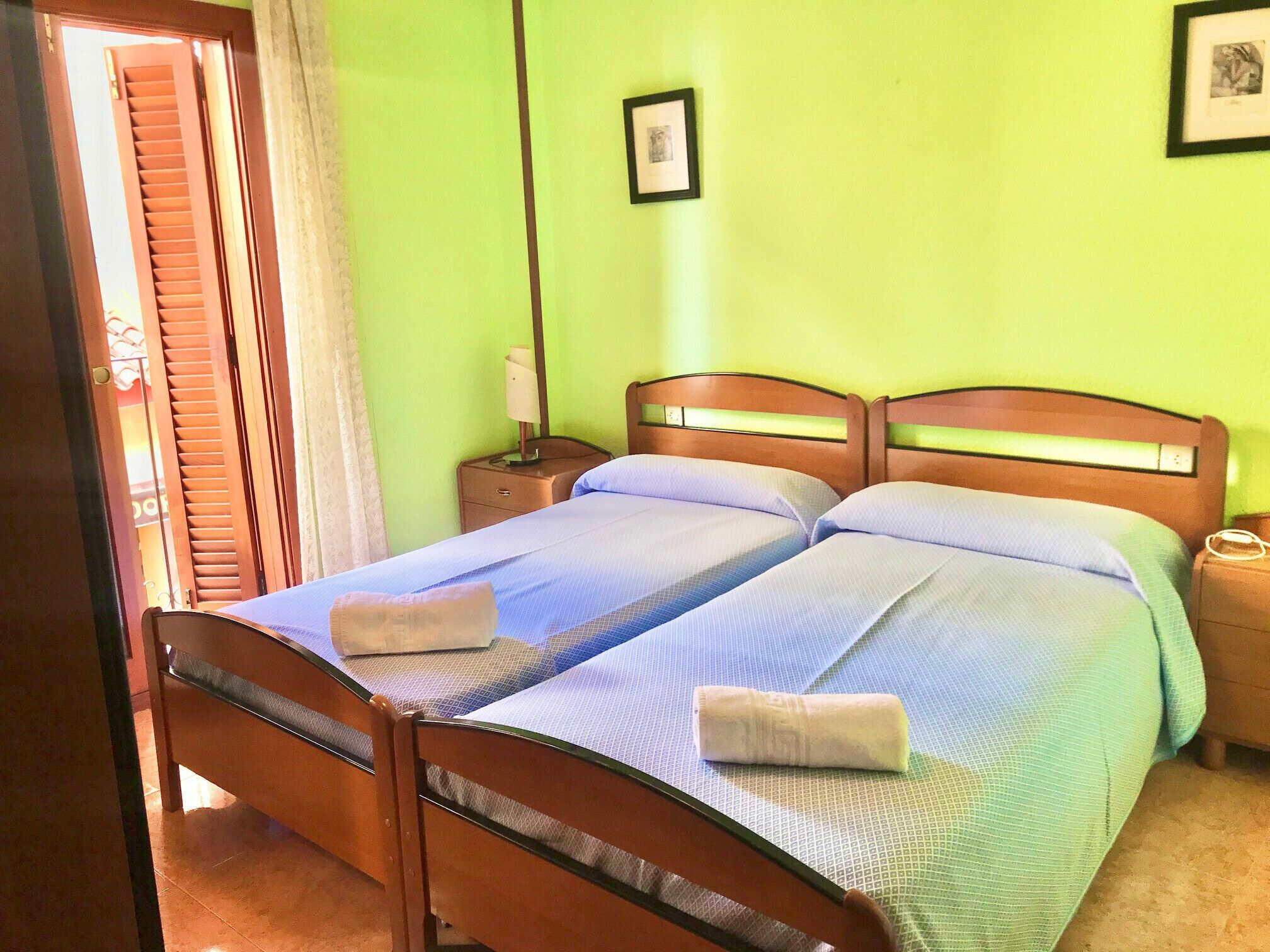 A double bed with two pillows and a folded towel on each side, a nightstand with a lamp, and a framed picture on the wall.