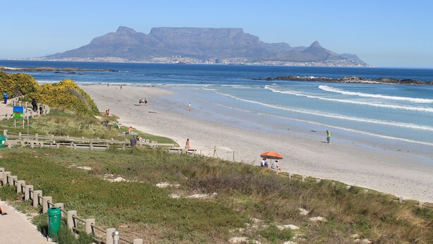 A beach with a view of a mountain in the background, a wooden fence, and a few people walking. - A REAL HOME AWAY FROM HOME EXPERIENCE.... (Cape Town)
