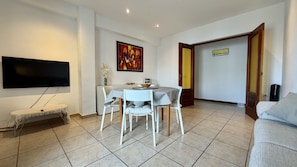 A living room with a TV, a dining table with chairs, a sofa, and a door leading to another room. - APARTMENT ELS PESCADORS 2-1 3D (L'Escala)