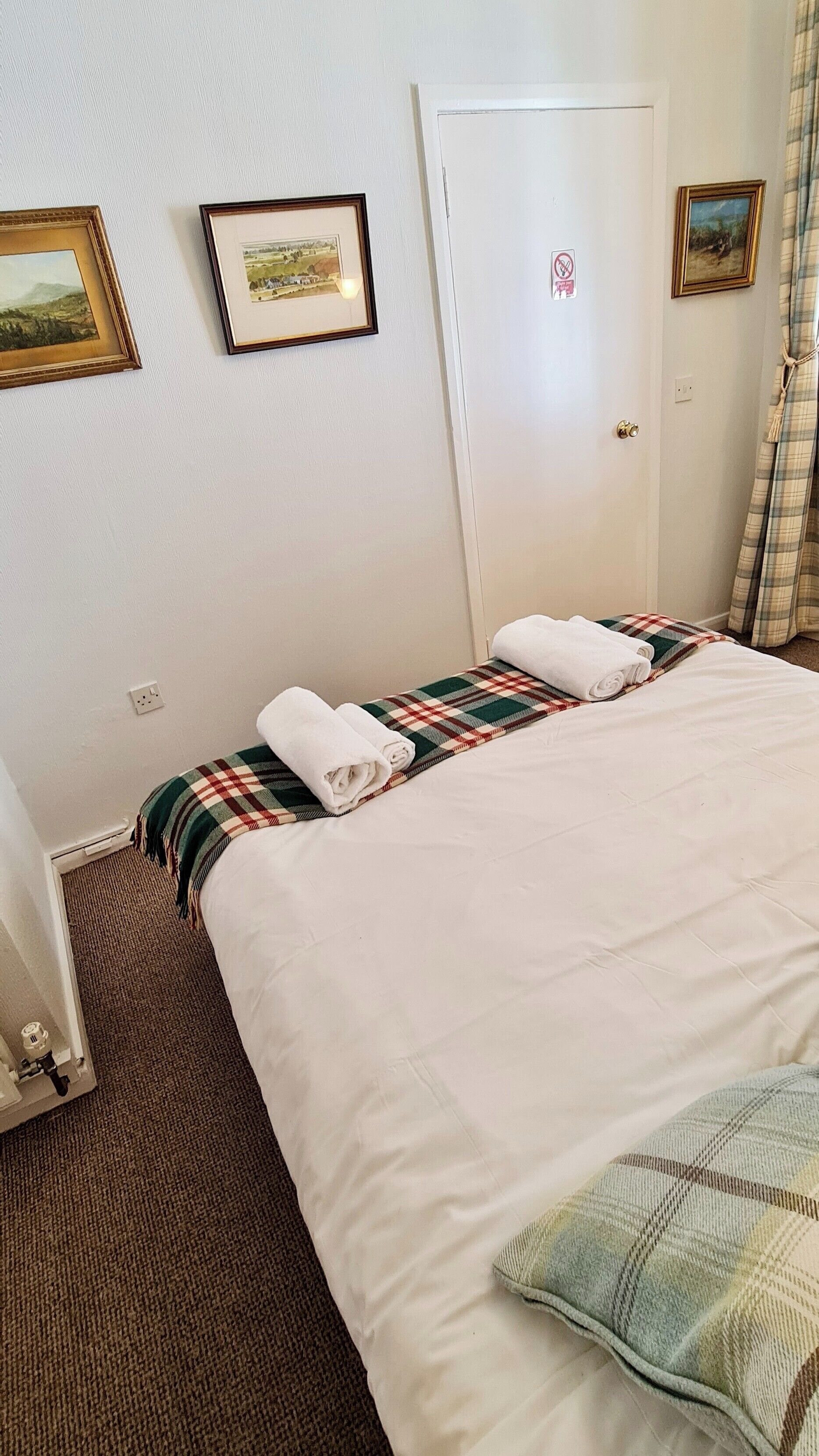Ein Hotelzimmer mit einem Bett, einer karierten Decke, zwei aufgerollten Handtüchern, einer Tür mit einem Rauchverbotsschild und zwei gerahmten Bildern an der Wand.