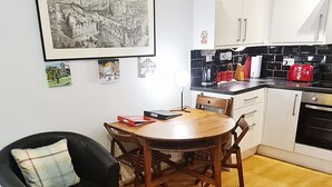 Un espacio habitable compacto con una mesa de comedor, un sillón de cuero negro, un tapete estampado, una cocina con armarios blancos y una imagen enmarcada de un paisaje urbano en la pared.