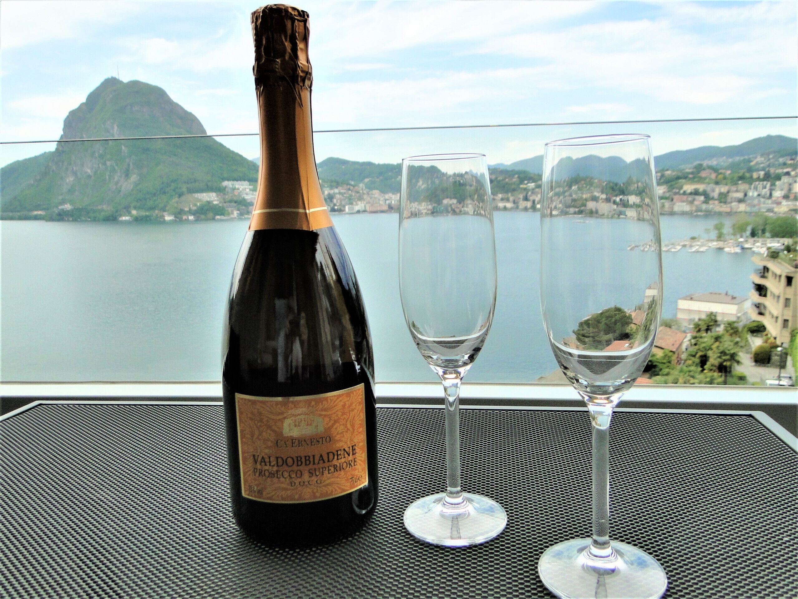 A bottle of Valdobbiadene Prosecco Superiore DOCG and two empty wine glasses on a table with a cityscape view.