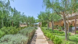 A pathway lined with greenery and trees, leading to a building with a stone facade and arched windows. - VILLA ES TRENC - SA SINIA VELLA (Campos)