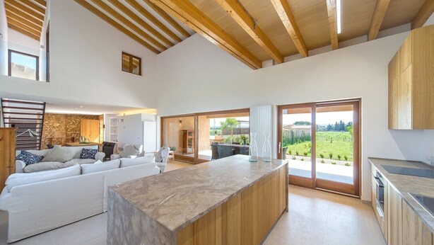 A spacious living area with a large wooden ceiling, a kitchen with wooden cabinets, and a sliding glass door leading to a garden. - VILLA ES TRENC - SA SINIA VELLA (Campos)