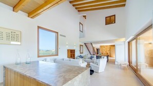 A spacious living area with a high ceiling, a central seating area with sofas, a wooden staircase, and a view through large windows. - VILLA ES TRENC - SA SINIA VELLA (Campos)
