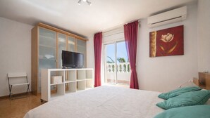 Ein Schlafzimmer mit einem Bett, einem Fernseher auf einer weißen Kommode, einem Bild an der Wand und Blick ins Freie.