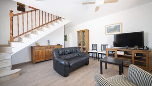 Ein Wohnzimmer mit Treppe, einer schwarzen Ledercouch, einem hundszahn-gemusterten Sessel, einem Holzschrank und einem Fernseher.