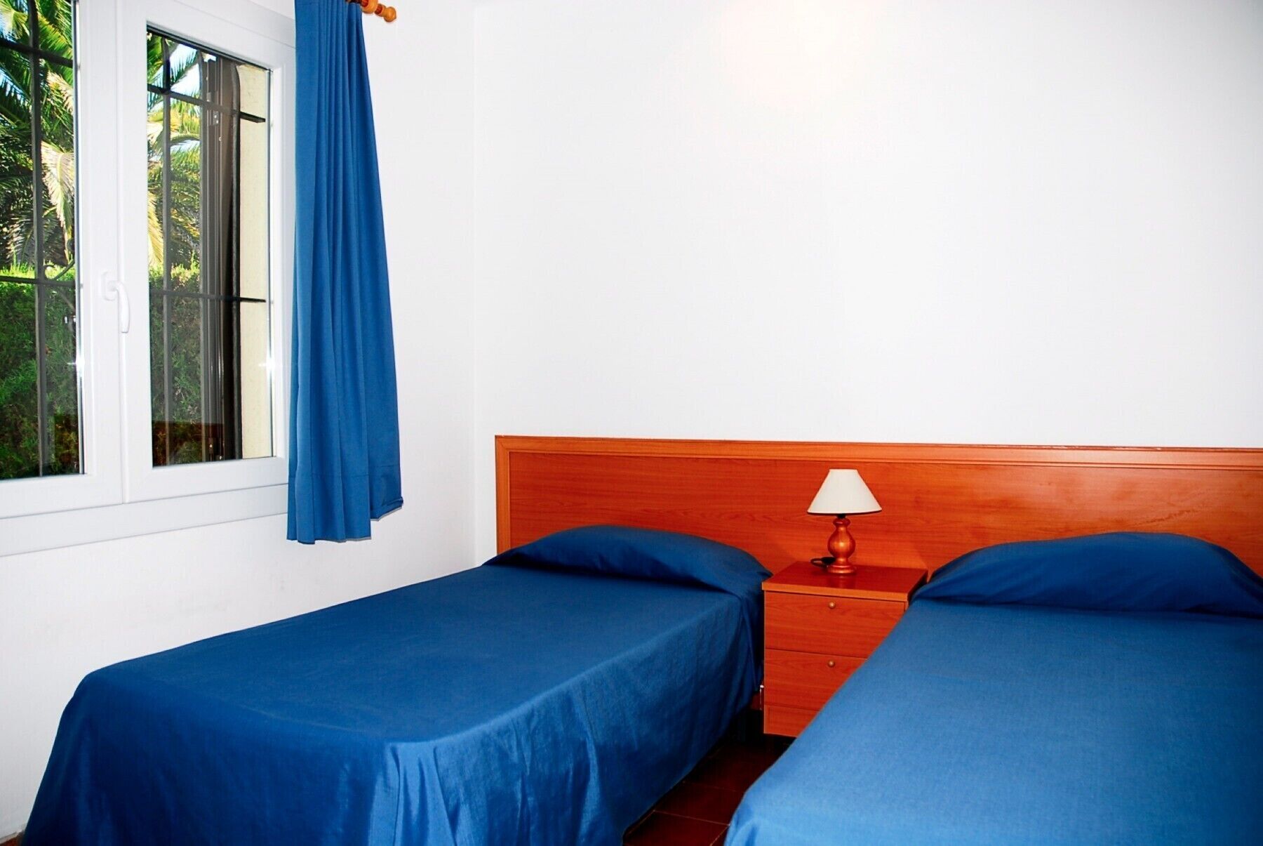 A room with two single beds, a wooden headboard, a nightstand with a lamp, and a window with a blue curtain.