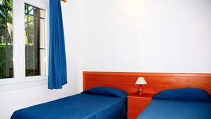 Une chambre avec deux lits simples, une tête de lit en bois, une table de chevet avec une lampe et une fenêtre avec un rideau bleu.