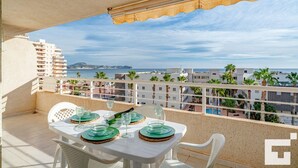 Varanda com mesa posta para dois, com vista para a costa, edifícios e palmeiras.