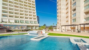 Uma piscina com uma pequena cascata, rodeada por um relvado bem cuidado e um edifício de vários andares com varandas.