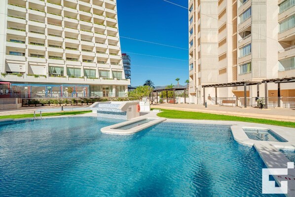 A swimming pool with a small waterfall feature, surrounded by a well-maintained lawn and a multi-story building with balconies.