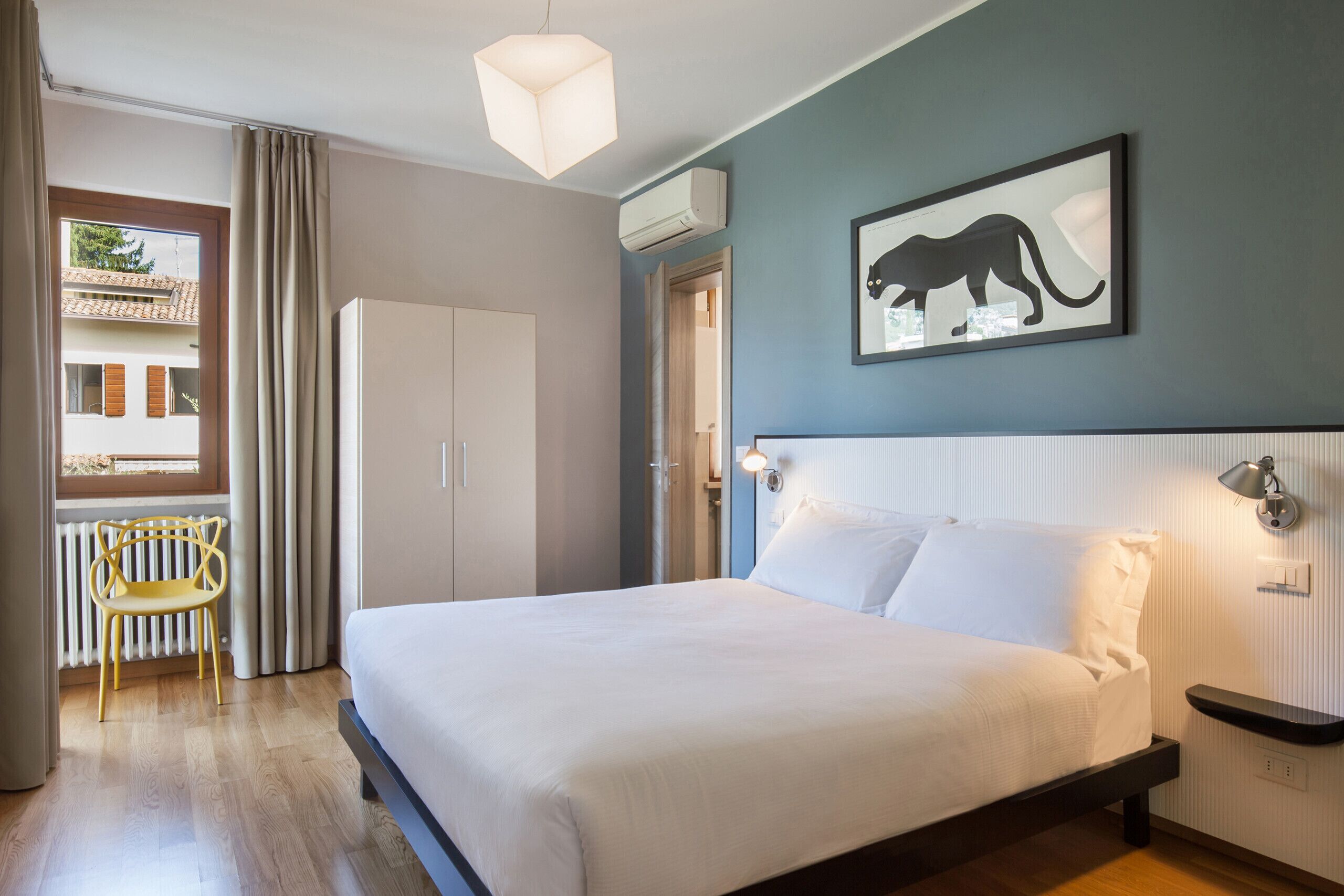 A bedroom with a bed, a chair, a window with curtains, a radiator, and a wall-mounted air conditioner.