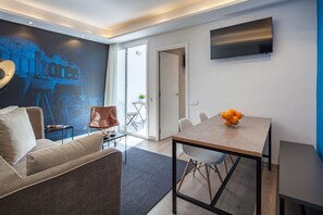 A modern living room with a sofa, a coffee table, a dining table with chairs, a TV mounted on the wall, and a large window.
