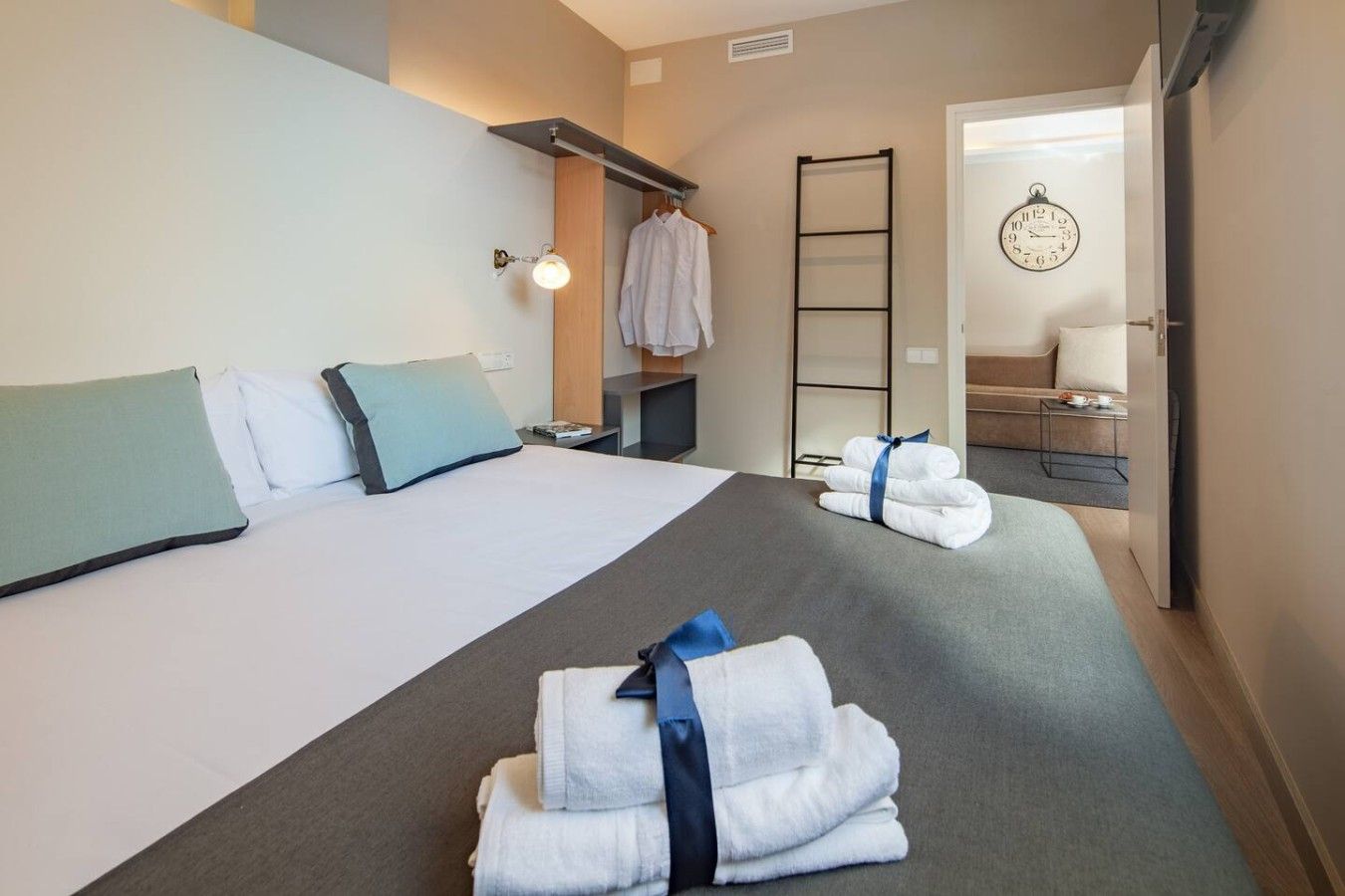 A hotel room with a bed, pillows, a bedside table, a lamp, a towel rack, a mirror, and a door leading to a bathroom.