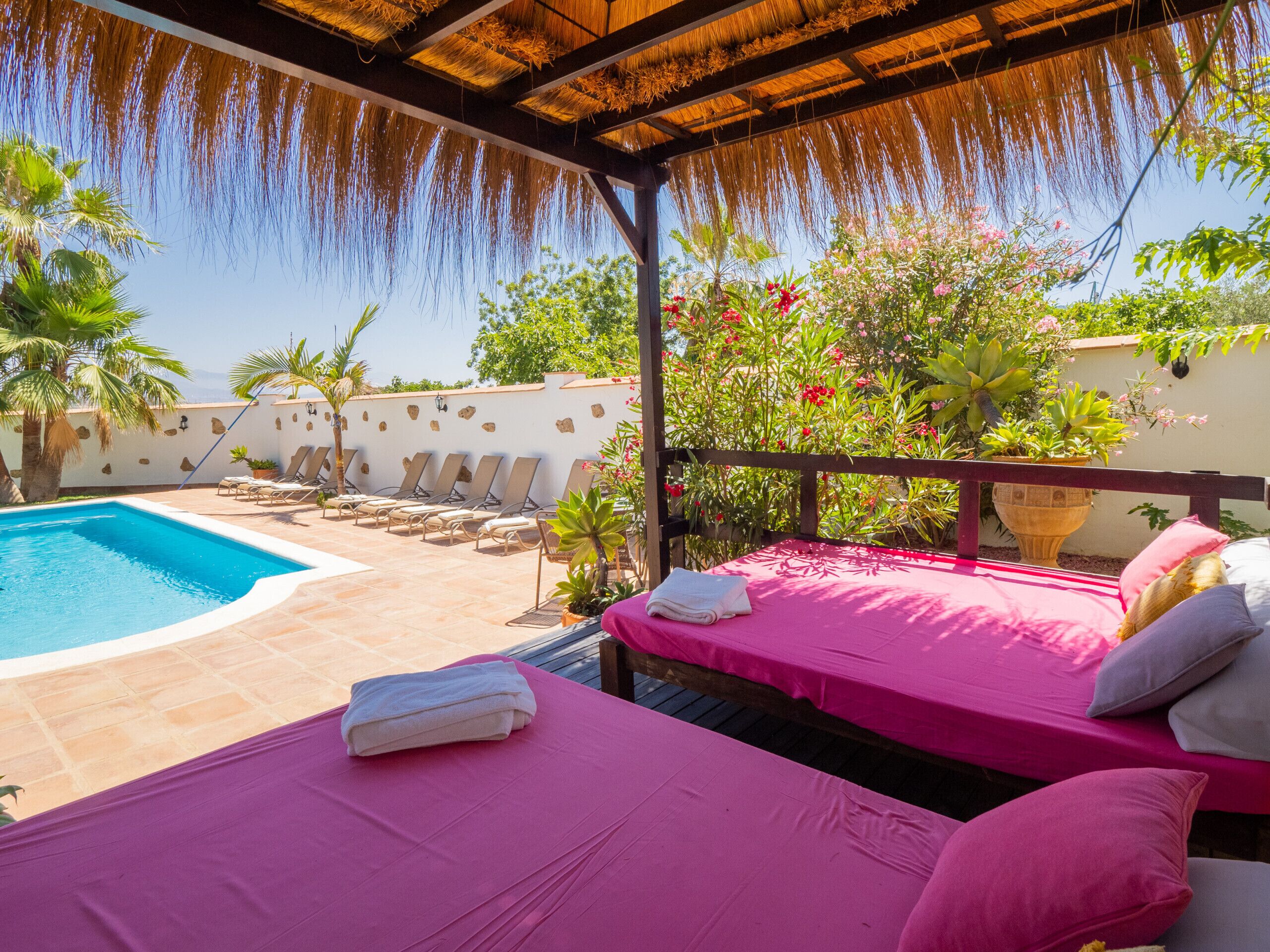 Un espace aménagé au bord de la piscine, doté d’un toit de chaume, de chaises longues et d’un lit avec une literie rose.