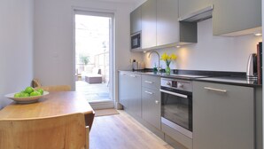 Une cuisine moderne avec des meubles gris, un four en acier inoxydable et un four à micro-ondes. On y trouve une table à manger en bois sur laquelle est posé un saladier rempli de pommes vertes.