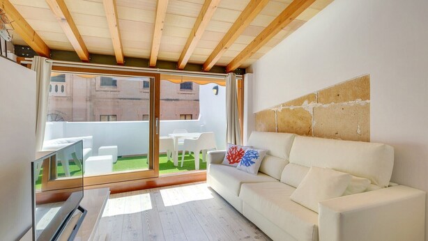 Een moderne woonkamer met een witte bank, een televisie en een schuifdeur naar een balkon met tuinmeubels.