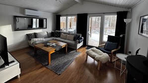 Een moderne woonkamer met een bank, salontafel en televisie. Er is een groot raam met gordijnen, een spiegel en een deur naar buiten.