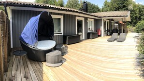 Een overdekt buitengebied met een houten terras, een overdekte zithoek met een tafel en stoelen, en een barbecue.