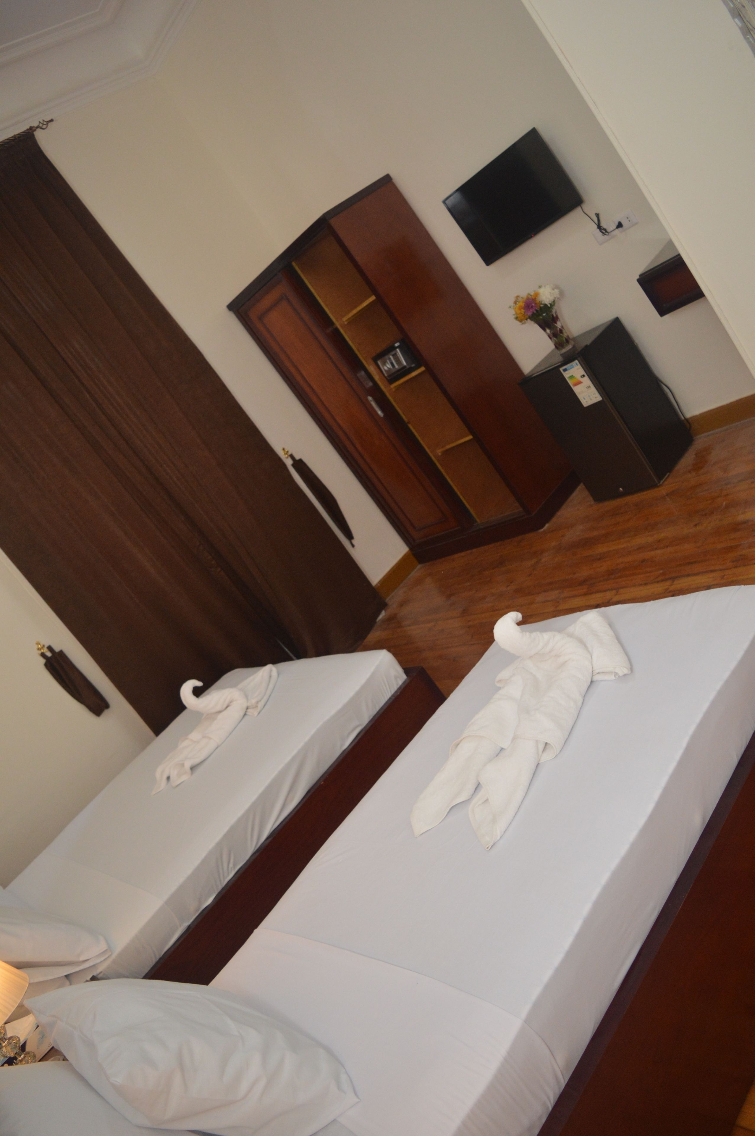 A hotel room with two beds, a wooden wardrobe, a flat-screen TV, and a small table with a vase of flowers.