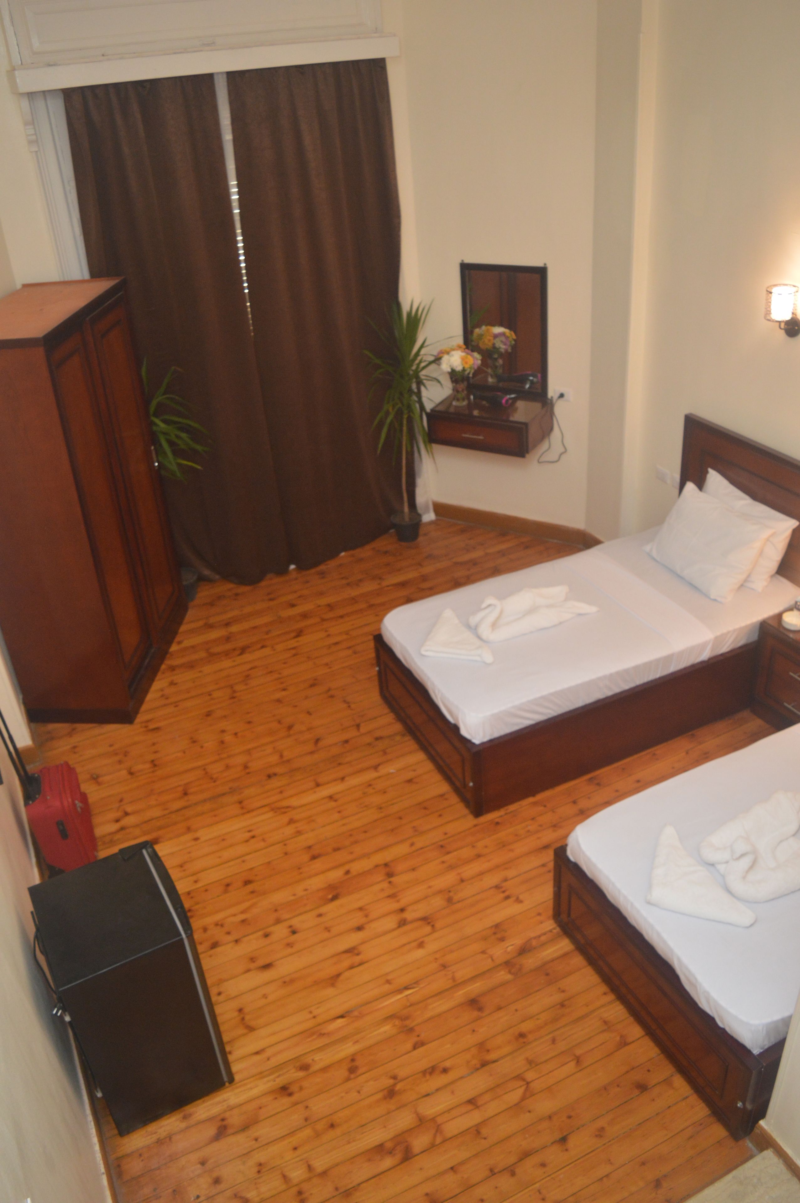 A room with a wooden floor, two beds, a wardrobe, a mirror, and a small table with a plant.