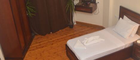 A room with a wooden floor, two beds, a wardrobe, a mirror, and a small table with a plant.