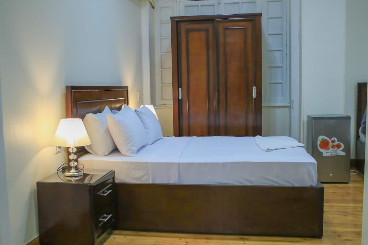 A bedroom with a bed, a nightstand, a lamp, a wardrobe, and a refrigerator.