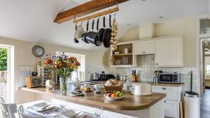 A modern kitchen with a wooden island, hanging pots, a clock, and a dining area set for a meal. - Downclose Piggeries, North Perrott , Dorset (North Perrott)