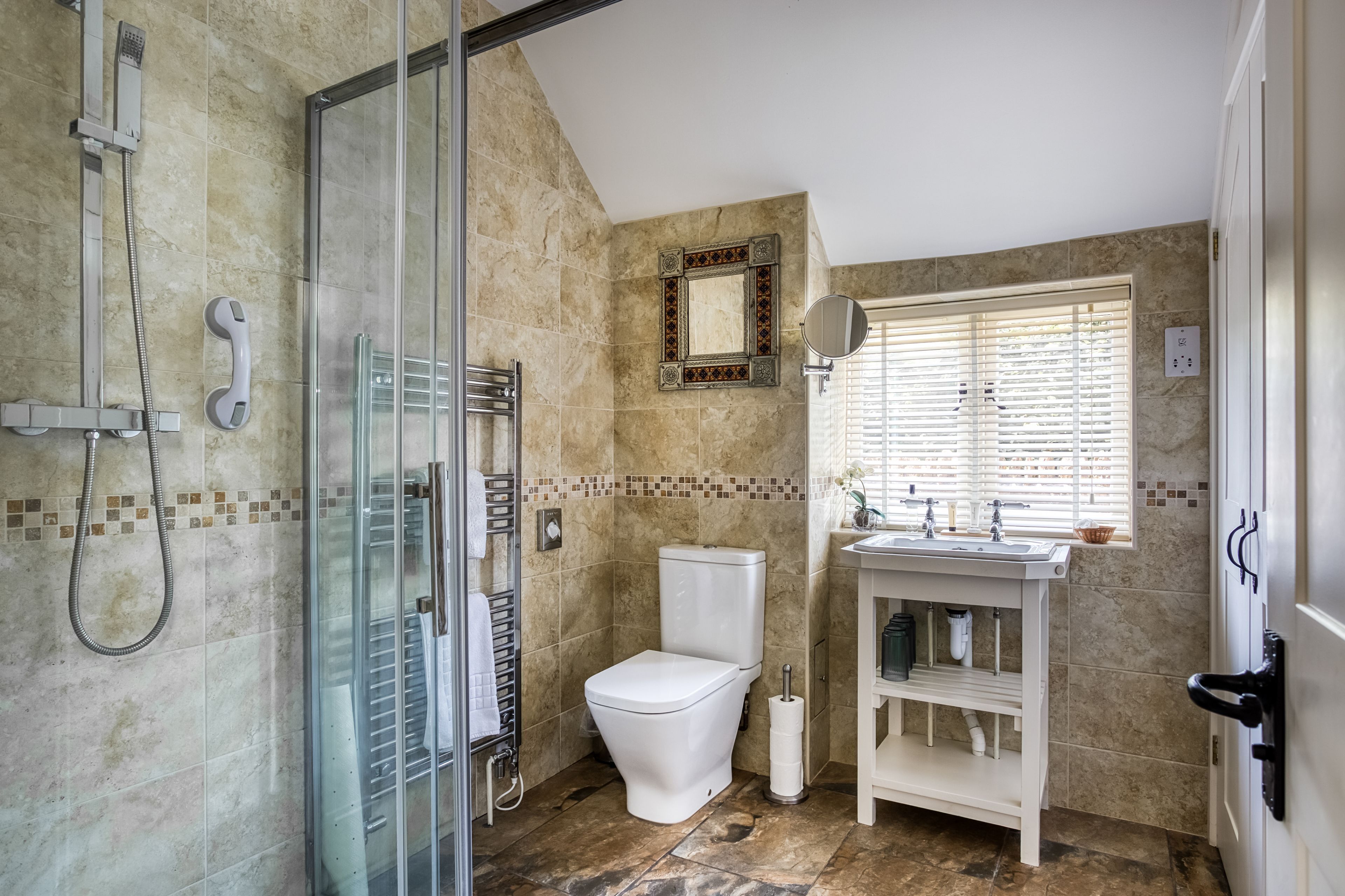 A bathroom with a glass shower enclosure, a toilet, a sink with a mirror, and a window with blinds.